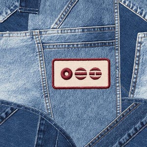 May include: Close-up of a patchwork denim design. The image features various shades of blue denim fabric pieces sewn together. A rectangular patch with the letters "OEE" in a maroon border is visible.