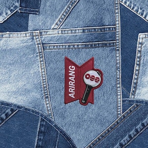 May include: A patchwork denim background with various shades of blue. A red patch with the word "ARIRANG" and a microphone design is sewn onto the denim. The microphone has the letters "OEE" on it.
