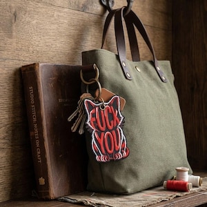 May include: A green canvas tote bag with brown leather handles hangs on a hook. A keychain with a red and black graphic that says "FUCK YOU" is attached to the bag. A leather-bound book and spools of thread are on a wooden shelf.