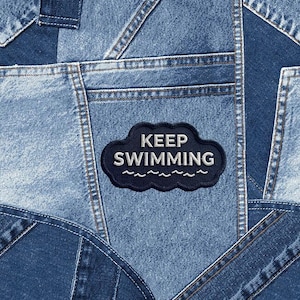May include: A patchwork denim design with various shades of blue. A dark blue cloud-shaped patch with the words "KEEP SWIMMING" in white is sewn onto the denim. The image showcases the texture and stitching details of the fabric.