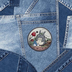 May include: Close-up of a denim patchwork design. A circular embroidered patch features a cartoon scene with a large gray creature, smaller blue creatures, and red mushrooms. The denim has various shades of blue.