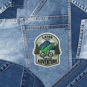 May include: A patchwork denim background with a patch that reads "Later Gator Time for Adventure." The patch features a green alligator riding a bicycle in front of a mountain scene. The denim has various shades of blue.