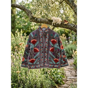 Puede incluir: Una chaqueta gris con bordados florales vibrantes en tonos rojo, naranja, azul cerceta y morado. La prenda cuenta con cuello mao, mangas largas y cierre frontal de botones, colgada en una percha de madera en un entorno natural.