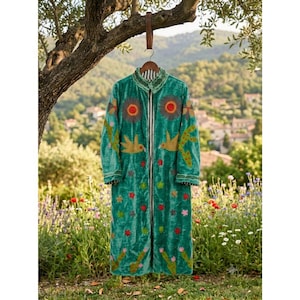 Pode incluir: Um casaco comprido de veludo verde-azulado com mangas compridas, decorado com motivos florais e de pássaros bordados em tons de laranja, amarelo e verde. A peça tem gola, fecho frontal com botões e um forro interior às riscas pretas e brancas.
