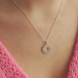 May include: A silver necklace with a circular pendant. The pendant features a detailed, openwork design. The necklace is displayed against a pink knitted garment.