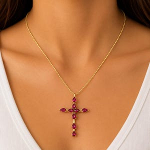 Puede incluir: Un collar de cadena de cuentas en tono dorado con un colgante de cruz. La cruz está construida con múltiples piedras preciosas de color rosa intenso en forma de lágrima engastadas en metal dorado, dispuestas en un patrón vertical y horizontal.