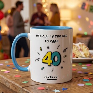 Peut inclure: Mug en céramique blanche avec une anse et un intérieur bleus, portant l'inscription "OFFICIALLY TOO OLD TO CARE." et le chiffre "40" en jaune et turquoise. Le mot "FINALLY." est écrit en dessous. Le mug est sur une table en bois avec des confettis.