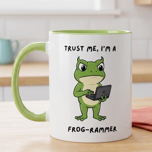 May include: A white ceramic mug with a light green handle and interior rim. The front features a cartoon green frog holding a small black laptop. Above the frog is the text TRUST ME, I'M A and below is the text FROG-RAMMER in black font.