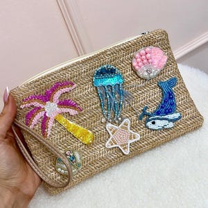 May include: A beige woven clutch bag with a zip and wrist strap. It features beaded sea-themed designs: a palm tree, jellyfish, starfish, whale, and seashell. The bag is held by a person.