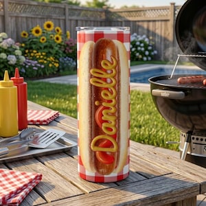 May include: A cylindrical tumbler featuring a red and white checkered pattern background with a central graphic of a hot dog. The name Danielle is written vertically in a yellow, mustard-like script font across the hot dog image.