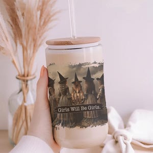 May include: A clear glass tumbler with a bamboo lid and a clear straw, featuring a printed graphic of five figures in witch hats standing in a circle around a candlelit table, with the text Girls Will Be Girls printed across the centre.