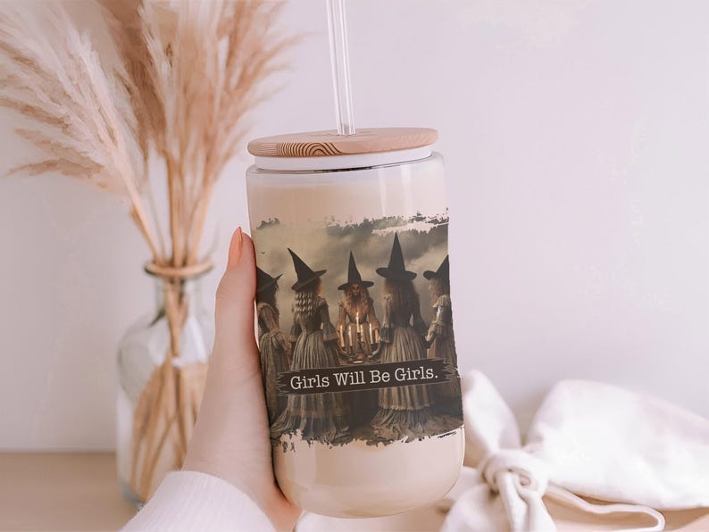 May include: A clear glass tumbler with a bamboo lid and a clear straw, featuring a printed graphic of five figures in witch hats standing in a circle around a candlelit table, with the text Girls Will Be Girls printed across the centre.
