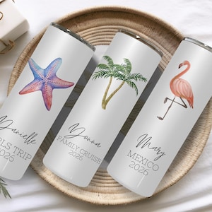 May include: Three white cylindrical tumblers with metal rims. The first features a purple starfish and the text Danielle GIRLS TRIP 2026. The second shows palm trees and Yonna FAMILY CRUISE 2026. The third displays a pink flamingo and Mary MEXICO 2026.