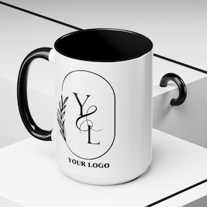May include: White ceramic mug with a black interior, handle, and rim. The mug features a black oval design with the letters "YL" and a leaf design, with the text "YOUR LOGO" below. A great gift for any occasion.