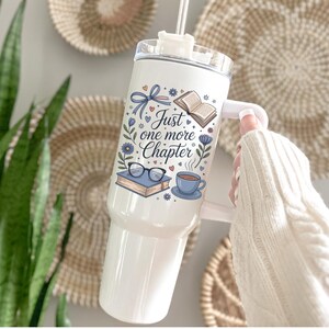 May include: White insulated tumbler with a handle and a straw. The tumbler features a floral design with the text "Just one more Chapter", an open book, glasses, and a cup of coffee. The tumbler is held by a person wearing a white sweater.
