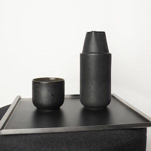 Könnte beinhalten: Eine schwarze Keramiktasse und ein hoher, schwarzer Keramikkrug stehen auf einem rechteckigen schwarzen Tablett mit silbernem Metallrand. Die Objekte befinden sich auf einer dunkelgrauen Oberfläche vor einem weißen Hintergrund. Die Tasse hat eine gesprenkelte Textur.