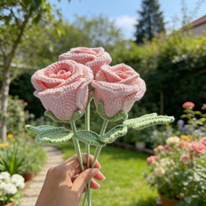 Puede incluir: Tres rosas de ganchillo hechas a mano en rosa suave, sostenidas contra un fondo de jardín borroso. Las rosas tienen pétalos detallados, tallos y hojas verdes. Un encantador arreglo floral hecho a mano.