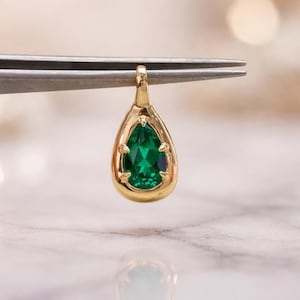 Puede incluir: Un colgante de oro con una piedra preciosa verde esmeralda en forma de pera. El colgante está sujeto por unas pinzas contra un fondo borroso. La piedra preciosa está facetada, reflejando la luz.