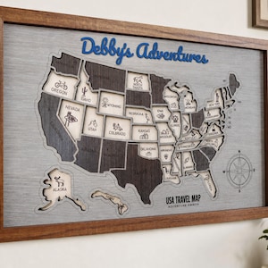 Könnte beinhalten: Eine gerahmte USA-Reisekarte aus Holz mit grauem Hintergrund, dunkelbraunen Staatsausschnitten und eingravierten Symbolen. Der Text Debby's Adventures steht in blauer Schrift oben, USA TRAVEL MAP ADVENTURE AWAITS steht unten rechts.