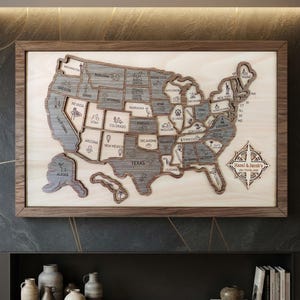 Pode incluir: Um mapa de parede de madeira dos Estados Unidos com estados em camadas cortados a laser em vários tons de cinza e madeira natural. O mapa inclui nomes de estados, pequenos ícones e uma rosa dos ventos com o texto Hazel & Jacob's USA Travel Map.