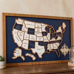 Op de afbeelding: Een ingelijste wandkaart van de Verenigde Staten met witte houten uitsparingen van staten met gegraveerde iconen en namen, gemonteerd op een donkerblauwe achtergrond. Een houten kompasroos ornament toont de tekst Hazel & Jacob's USA Travels, 2023.