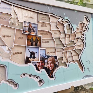 May include: A wooden United States map with individual state cutouts, each with a photo. The map is framed in white and mounted on a light blue background. The photos show people and landscapes.