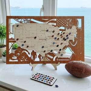 May include: A wooden map of the United States with football-shaped pins marking locations. The map is laser-cut with football player silhouettes and an American flag design. A brown football and a tray of pins are also present.