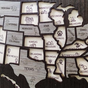 May include: A wooden wall map of the United States featuring laser-cut state borders. Each state is labelled with its name and includes a small engraved icon representing local landmarks or culture, such as a bison in Montana and a rocket in Alabama.
