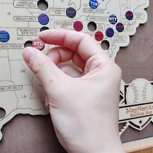 May include: Wooden baseball stadium map with colorful pins. A hand holds a pin labeled "STL" for St. Louis. The map includes locations like Yankee Stadium, Wrigley Field, and Fenway Park. The map is a decorative item.