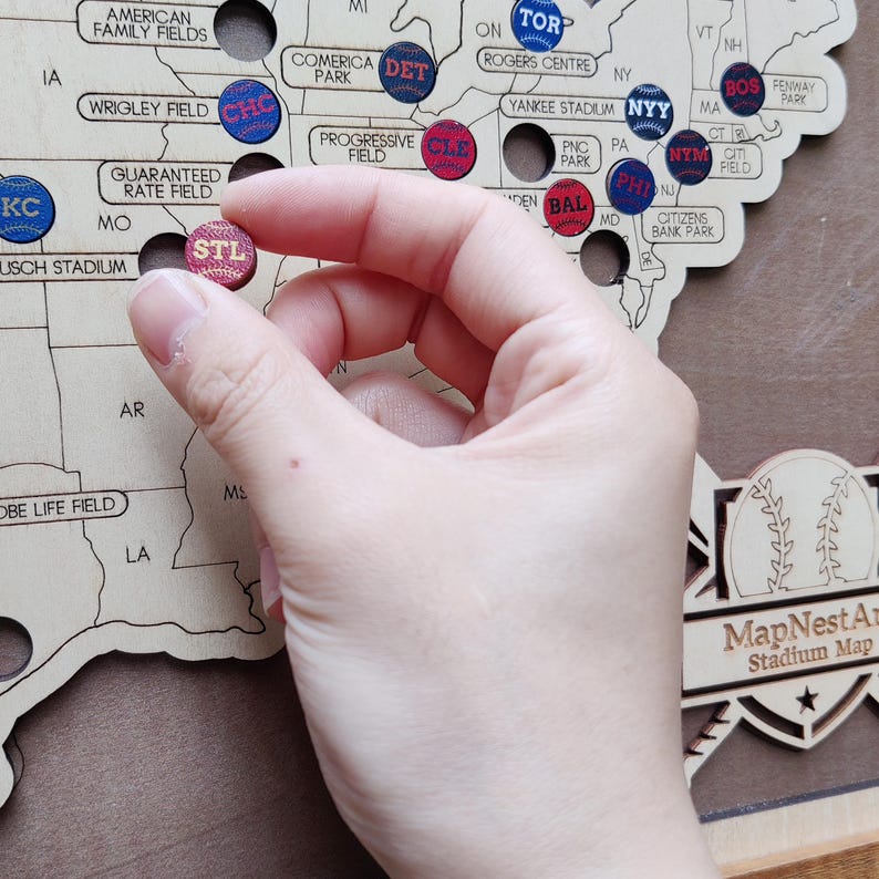 May include: Wooden baseball stadium map with colorful pins. A hand holds a pin labeled "STL" for St. Louis. The map includes locations like Yankee Stadium, Wrigley Field, and Fenway Park. The map is a decorative item.