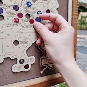 May include: A wooden map of the United States with baseball stadium markers. A hand is placing a red and blue marker labeled "PHI" on the map. Other markers include "DET", "CLE", "CIN", "BAL", and "MIA".