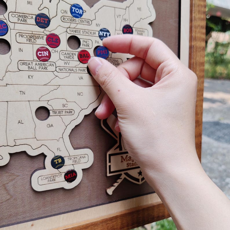 May include: A wooden map of the United States with baseball stadium markers. A hand is placing a red and blue marker labeled "PHI" on the map. Other markers include "DET", "CLE", "CIN", "BAL", and "MIA".