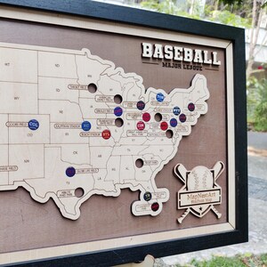 May include: Wooden baseball stadium map with a black frame. The map features a laser-cut design of the United States with stadium locations marked. The text "BASEBALL MAJOR LEAGUE" is at the top. The MapNestArt logo is at the bottom.
