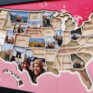 May include: A wooden wall map of the United States featuring laser-cut state borders and engraved state names. Each state section contains a photograph of a couple in various locations. The map is set against a bright pink background with grid lines.
