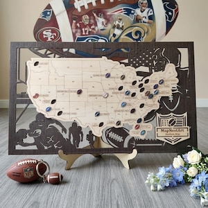 May include: A wooden stadium map of the United States with football-shaped pins marking locations. The map is framed in dark wood with football player silhouettes. A football keychain and a decorative football are in the foreground.
