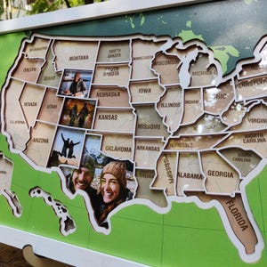 May include: A wooden wall map of the United States featuring laser-cut state borders and names. The map includes several rectangular photo inserts placed over specific states, showing two people in various outdoor settings and travel locations.