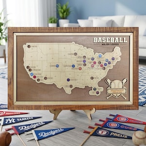 May include: A wooden baseball stadium map of the United States, with team markers. The map is framed in a light brown wood and includes the text "BASEBALL MAJOR LEAGUE". Several team pennants and a baseball are in the foreground.