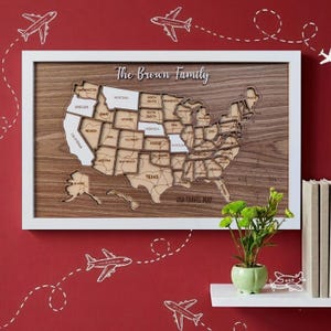 May include: Wooden USA travel map in a white frame, with state outlines and the text "The Brown Family". The map is on a red wall with airplane illustrations. A small green plant in a pot sits on a white shelf.