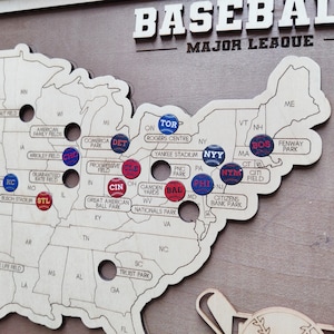 May include: Wooden baseball map of the United States with team pins. The word "BASEBALL" is at the top. Team pins include "TOR", "NYY", "BOS", "STL", "CHC", "CIN", "CLE", "DET", "PHI", "BAL", "NYM", and "KC".