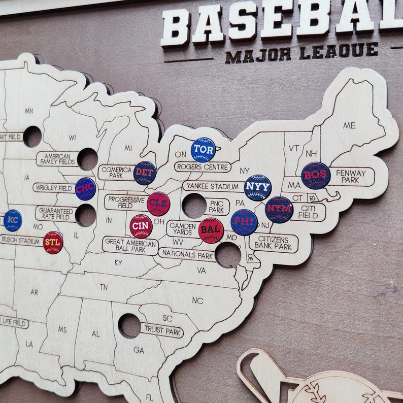 May include: Wooden baseball map of the United States with team pins. The word "BASEBALL" is at the top. Team pins include "TOR", "NYY", "BOS", "STL", "CHC", "CIN", "CLE", "DET", "PHI", "BAL", "NYM", and "KC".