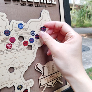 May include: A wooden Major League Baseball stadium map with team logo pins. The map is a light wood color, with the United States outlined. A hand is placing a Boston Red Sox pin on the map. The map includes the text "MapNestArt Stadium Map".