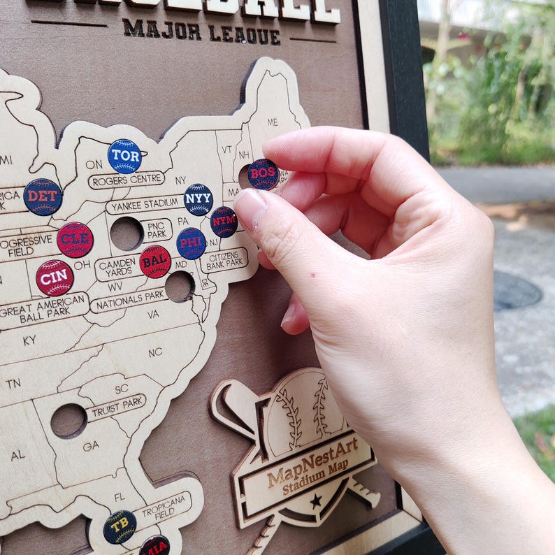 May include: A wooden Major League Baseball stadium map with team logo pins. The map is a light wood color, with the United States outlined. A hand is placing a Boston Red Sox pin on the map. The map includes the text "MapNestArt Stadium Map".