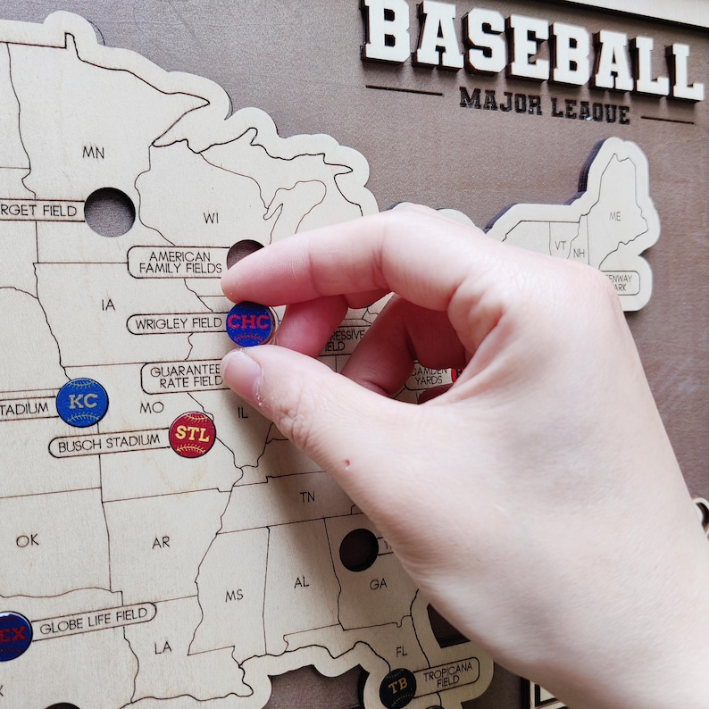 May include: Wooden baseball map with Major League Baseball text. The map features states with baseball stadium pins. A hand is placing a blue and red pin labeled "CHC" on the map.