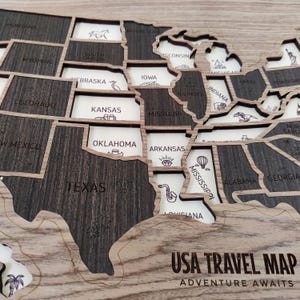 Op de afbeelding: Een houten USA reiskaart met lasergesneden staatsgrenzen. Donkere houten stukken vertegenwoordigen de staten, terwijl lichte houten stukken staatsnamen en gegraveerde iconen bevatten zoals een diamant, saxofoon en palmbomen. De tekst USA TRAVEL MAP ADVENTURE AWAITS is zichtbaar.