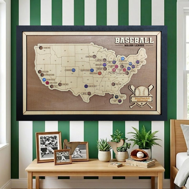 May include: A framed wooden baseball map of the United States, with team logos marking locations. The map is set against a green and white striped wall. The word "BASEBALL" is at the top. A baseball and bat graphic is on the right. Additional framed photos and plants are on a wooden table.