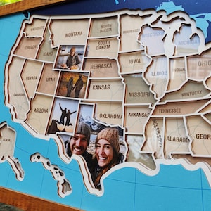 May include: A wooden wall map of the United States featuring laser-cut state borders and labels. The map includes integrated photo frames displaying personal images of people in various locations, set against a blue background with a wooden frame.