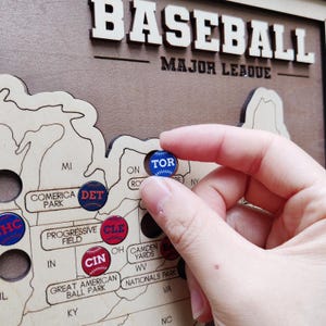 May include: A wooden baseball map with the word "BASEBALL" at the top. The map has cutouts of the United States and includes team markers. A hand holds a blue and white "TOR" marker, representing the Toronto Blue Jays.