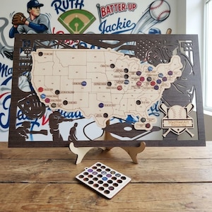 May include: A wooden baseball stadium map of the United States, with cut-out state shapes and stadium markers. The map is framed in dark brown wood and includes a separate stand and a small tray of stadium markers. The background features baseball-themed artwork.