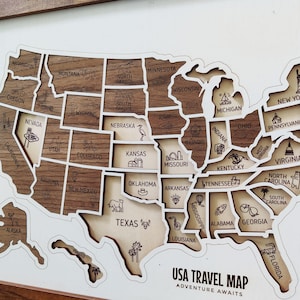 Puede incluir: Un mapa de viaje de EE. UU. de madera con formas de estados cortadas con láser en tonos de madera claros y oscuros alternos. Cada estado está etiquetado con su nombre y un pequeño icono grabado que representa un hito o símbolo local. El texto USA TRAVEL MAP ADVENTURE AWAITS está impreso en la esquina inferior derecha.