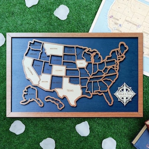 May include: A wooden map of the United States with state outlines. The map is set against a dark blue background and framed in a light brown wood. States are labeled with their names. The map includes a compass rose with the names "Patrick & Fiona" and "USA TRAVEL MAP".